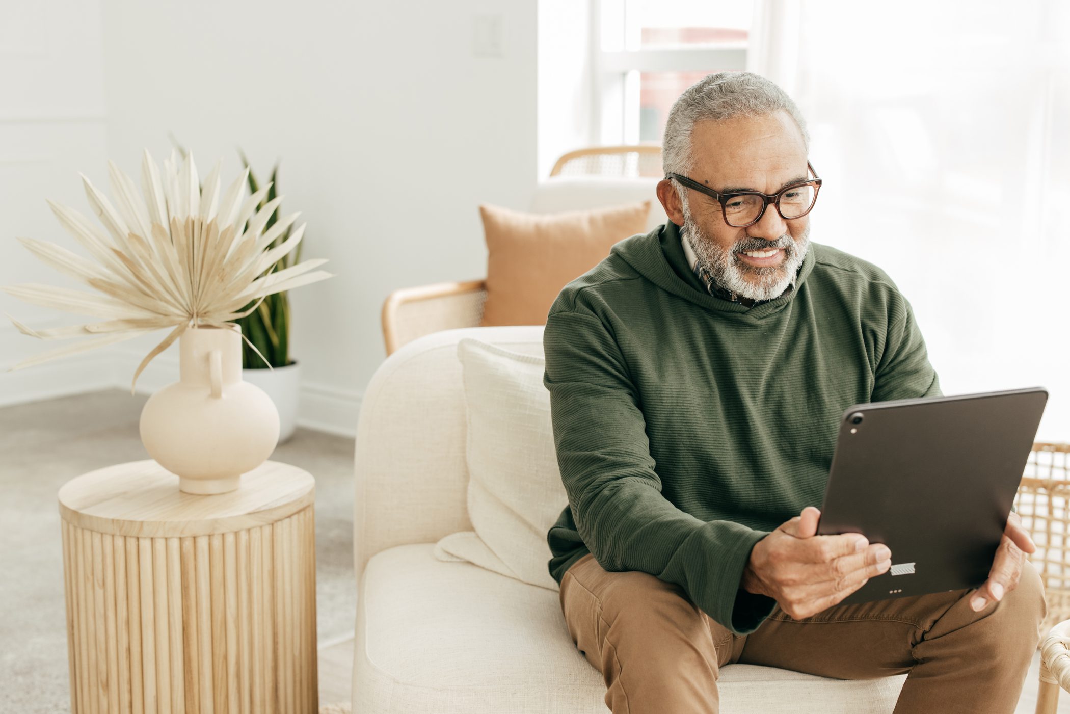 A mature sits on a comfortable chair and holds up his tablet.