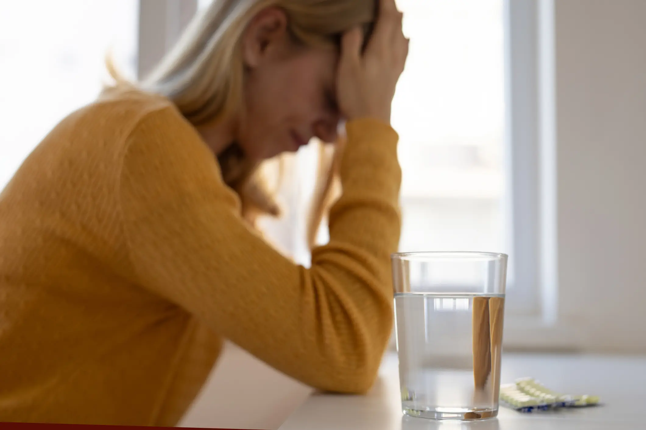 Struggling woman sits at a table, a glass of water and prescription medication in front of her.